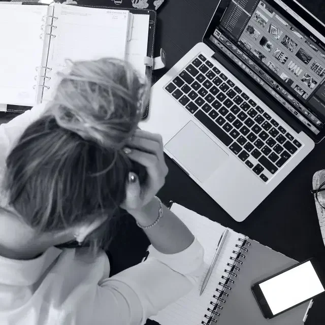 Frau sitzt verzweifelt mit Händen am Kopf über ihren Laptop, Kalender  und leeren Blocl.
